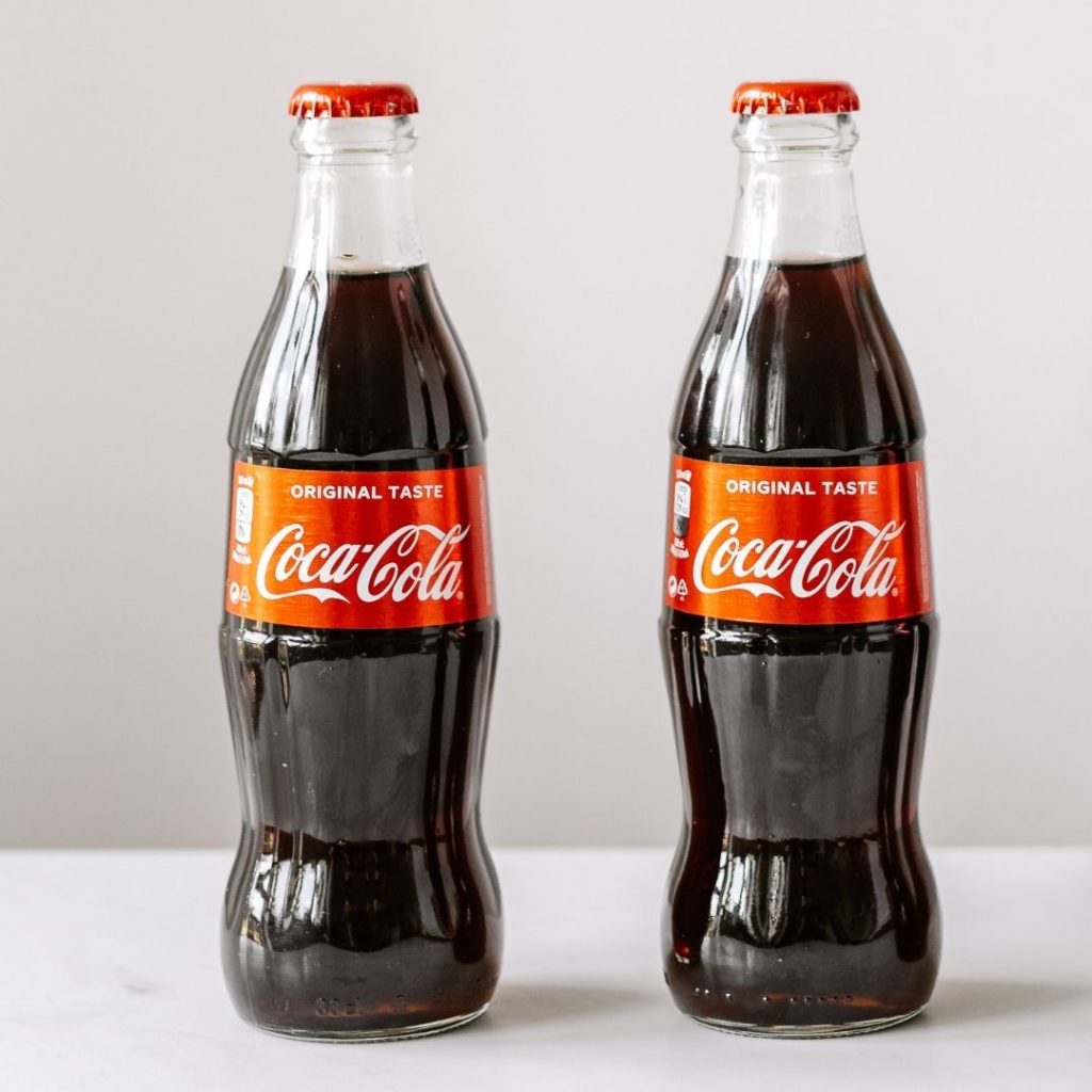 2 small bottles of coke on a granite bench with a plain offwhite background.